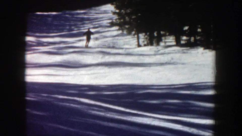 1961 Aspen home-movie frame with a distant skier moving across a bright snow-covered slope near evergreen trees.