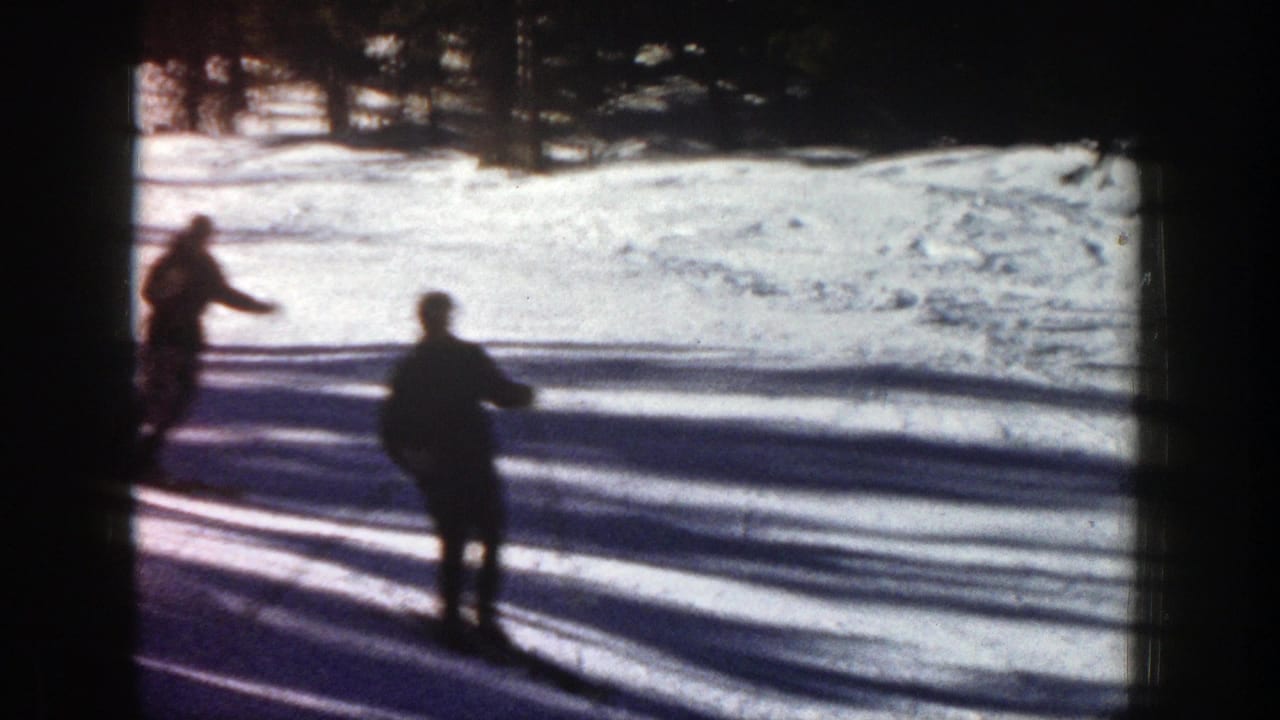 Two silhouetted skiers cross a snowy slope near a dark tree line.