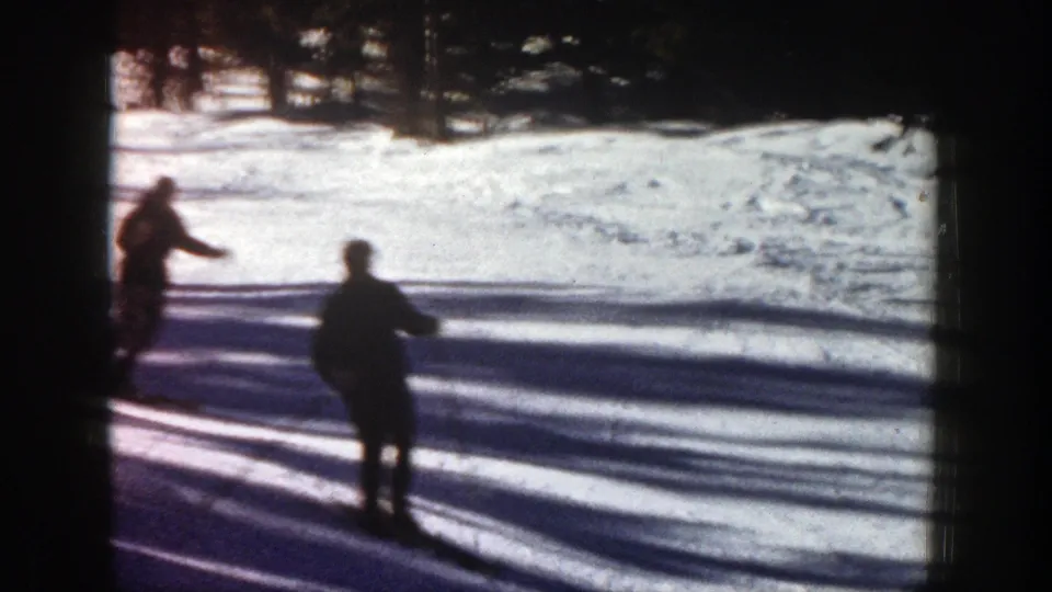 1961 Aspen home-movie frame with two silhouetted skiers on a bright snowy slope, framed by dark trees and long shadows.