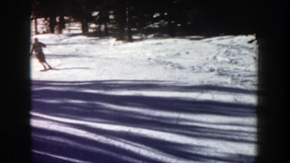 1961 Aspen home-movie frame with a skier entering from the left against a bright snowy slope and dark trees.