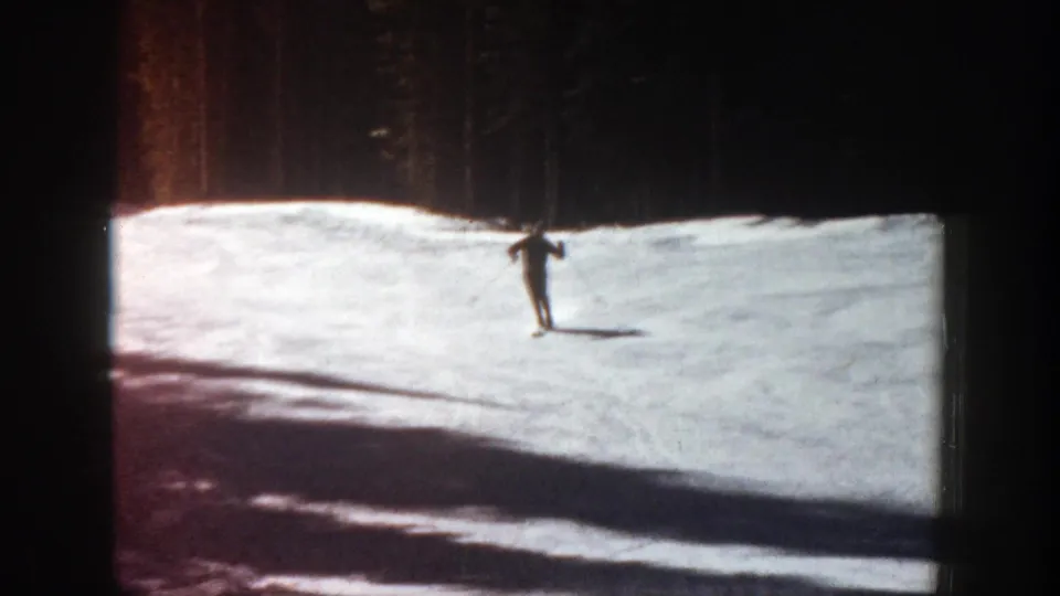 A distant skier crosses a bright snowfield beneath dark trees in 1961 Aspen home-movie footage.