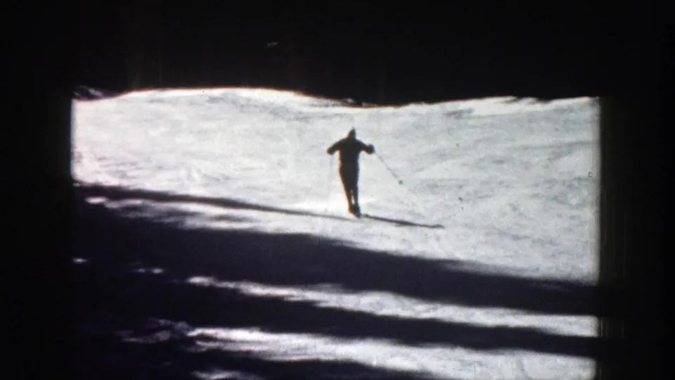The skier continues across the snow at a distance, framed by heavy shadows and wooded terrain in Aspen, Colorado.