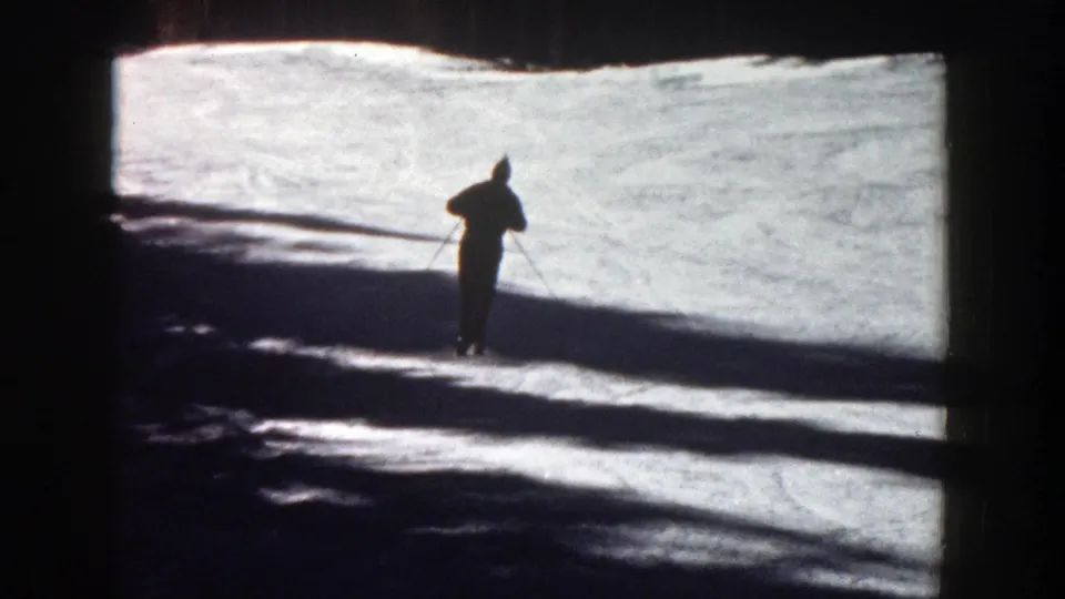 A lone skier stands or glides near the center of a sunlit Aspen snow slope, partly silhouetted by shadow.