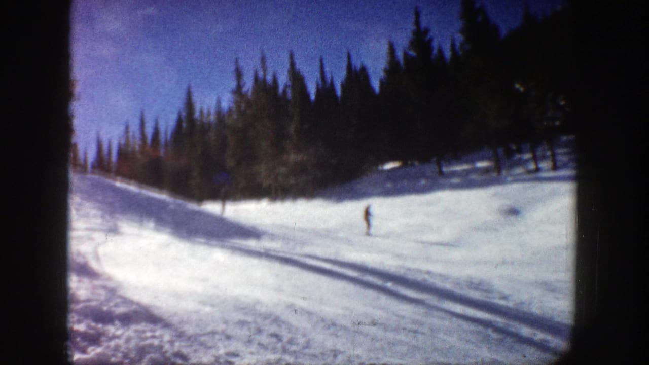 Snowy ski slope with evergreen trees and a distant skier under a blue sky.