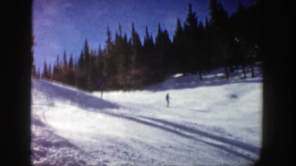 1961 Aspen-area 8mm frame showing a snowy slope, dark treeline, long shadows, and a small skier crossing the run.