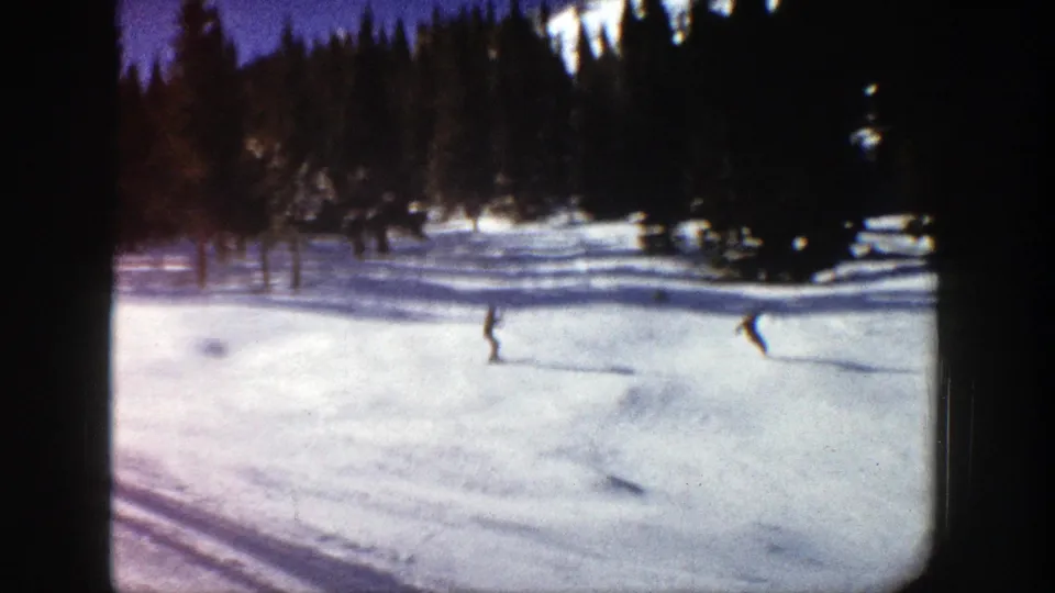 1961 Aspen home-movie frame showing two skiers spaced across a gentle snowy slope near a dense evergreen backdrop.