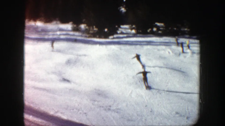 1961 Aspen home-movie frame showing skiers on a sunlit snowy slope with long shadows and tree cover above.