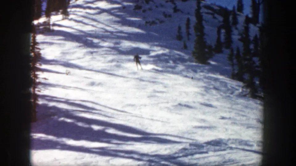 1961 Aspen ski scene with a distant skier on a sunlit slope, pine trees at the side, and strong shadows across the snow.