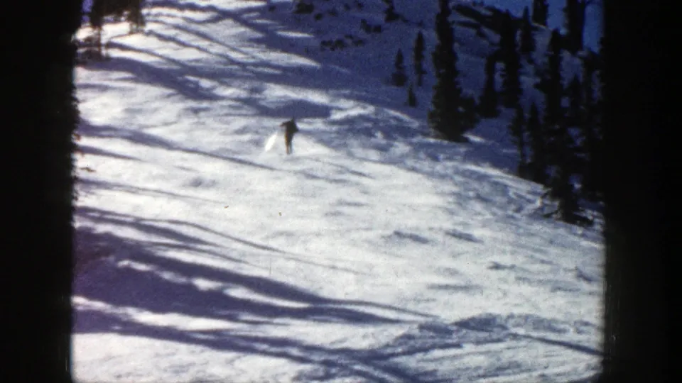 In Aspen, Colorado in 1961, a distant skier moves downhill across sunlit snow with dark evergreen trees nearby.