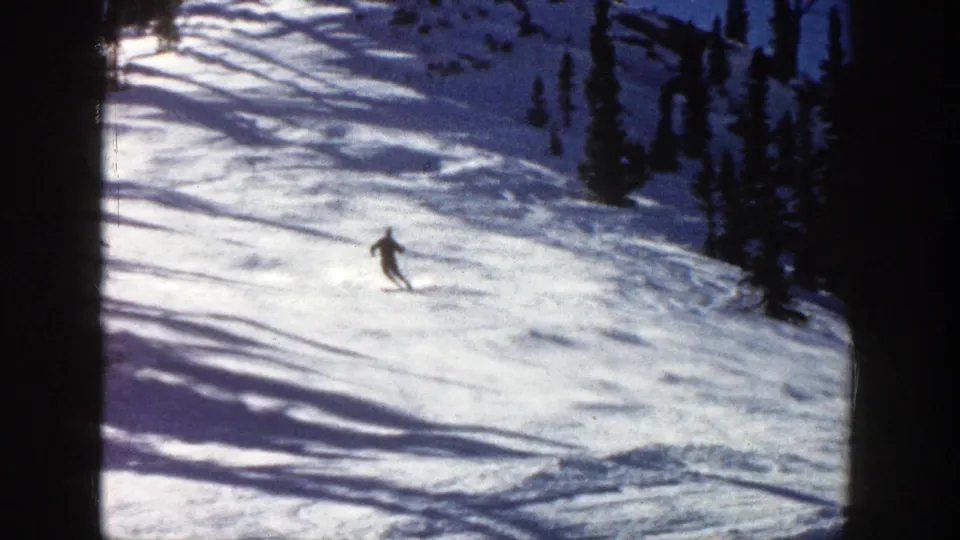 A skier in dark winter clothing descends the sunlit Aspen slope, with long shadows stretching across the snow.