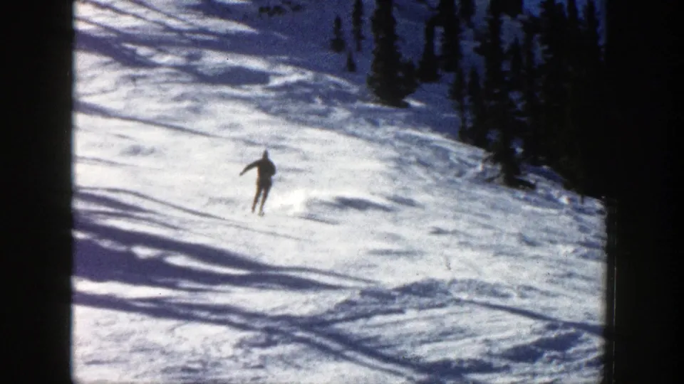 A skier in dark clothing and a blue hat descends a sunny snowy slope in Aspen, Colorado, in 1961 home-movie footage.