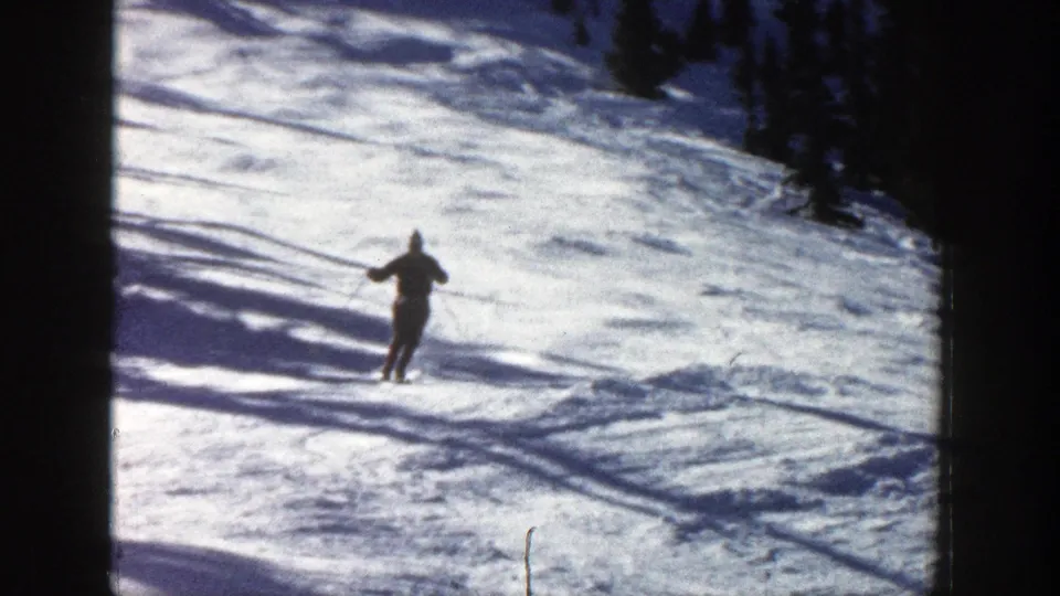 A skier in a dark outfit and blue hat comes closer while descending the snowy Aspen slope in 1961 8mm footage.