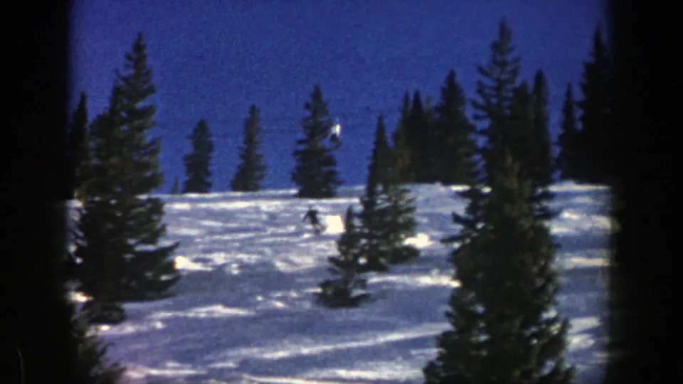 1961 Aspen 8mm frame of a skier moving across a snow-covered hillside between tall evergreen trees.