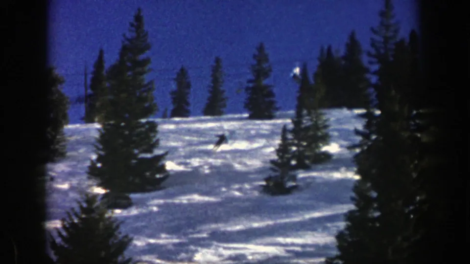 Final 1961 Aspen frame showing a distant skier descending a snowy run between evergreen trees under a blue sky.