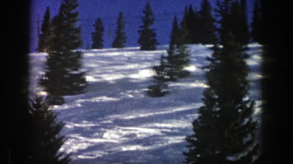 The skier continues downhill across a bright, tree-dotted Aspen snowfield, partly small and distant within the 8mm frame.