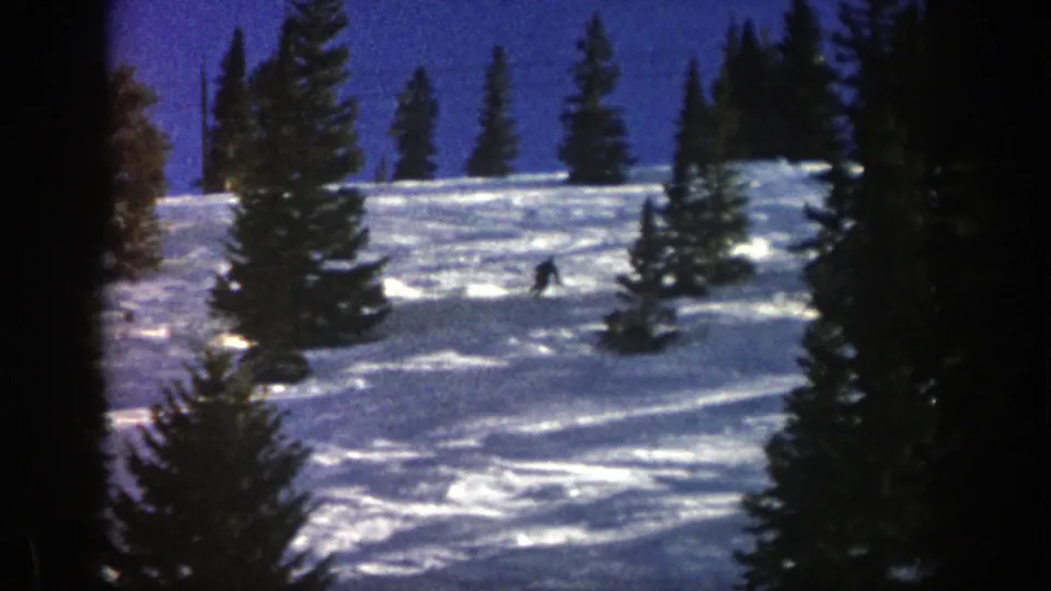 A dark-clad skier appears lower on the wooded Aspen slope, turning across sunlit snow in vintage 8mm footage from 1961.
