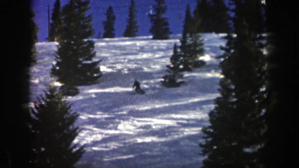 A small skier crosses the snow-covered Aspen hillside, partly surrounded by dark evergreens in 1961 home-movie footage.