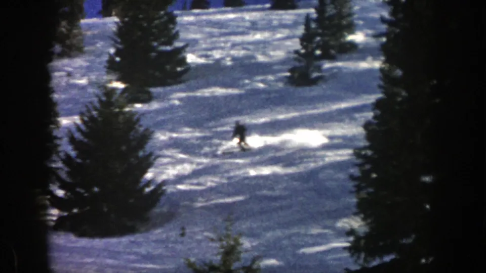 The lone skier continues across a bright Aspen snowfield, with evergreens and long shadows shaping the hillside view.