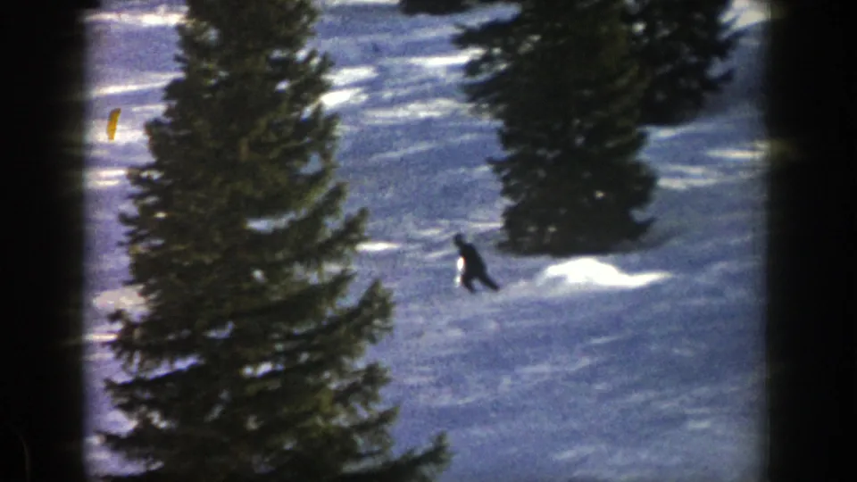 1961 Aspen home-movie frame with a distant dark-clad skier descending across a snowy slope between tall evergreen trees.