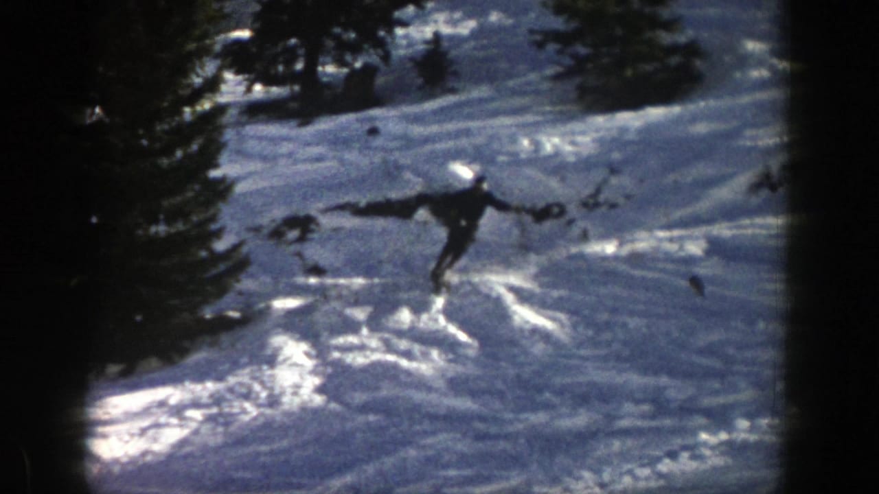 In 1961 Aspen footage, a distant skier in dark winter gear cuts across sunlit snow on a wooded mountain slope.