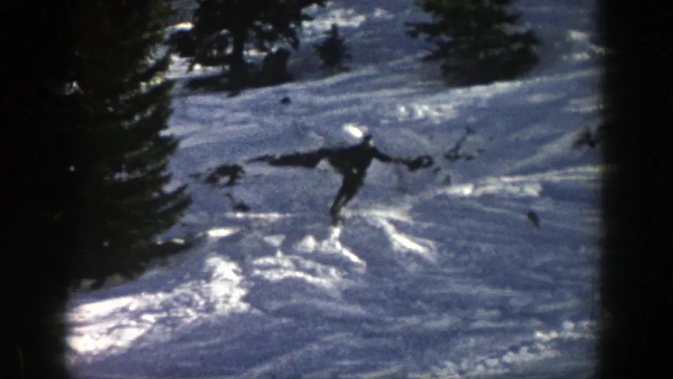 In 1961 Aspen footage, a distant skier in dark winter gear cuts across sunlit snow on a wooded mountain slope.