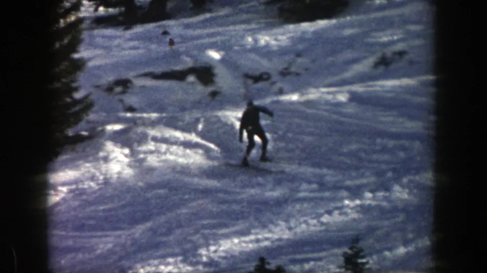 The skier continues downhill through a forested Aspen, Colorado slope, seen as a dark figure against reflective snow.
