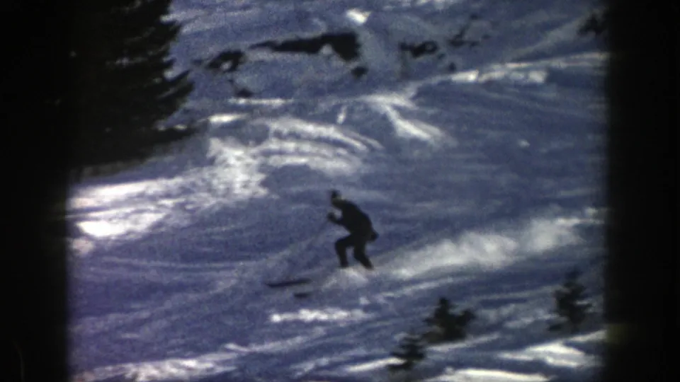 A distant skier crouches into a turn on tracked snow, with evergreen trees marking the edge of the Aspen slope.