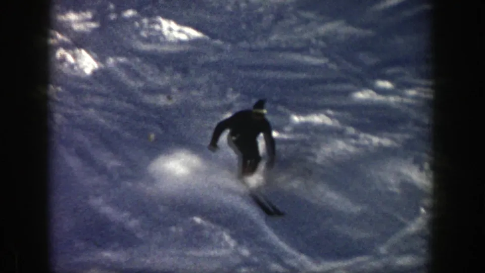 A skier in a dark jacket and knit cap carves through snowy moguls in the Aspen, Colorado home-movie footage.