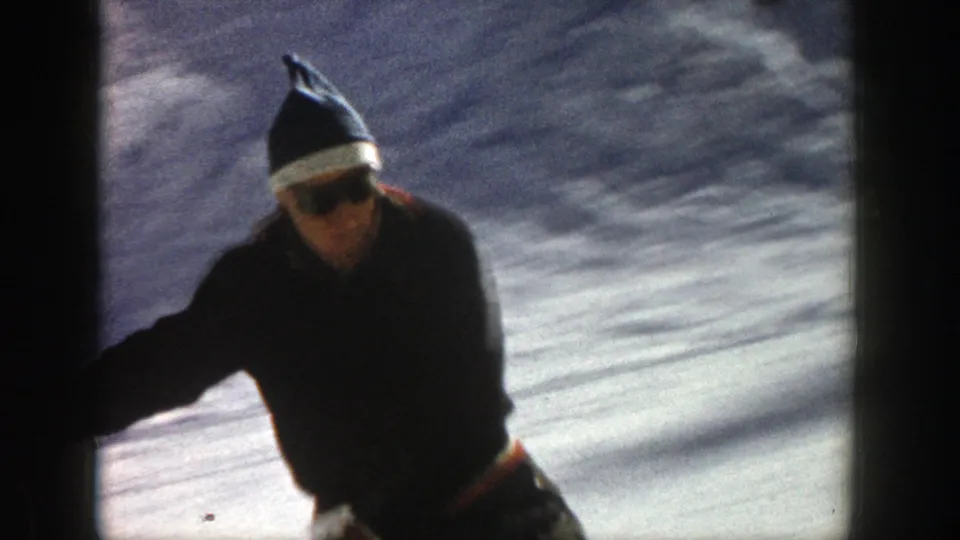 A close home-movie frame shows the skier passing near the camera in Aspen, with sunglasses, a knit cap, and snowy slope behind him.