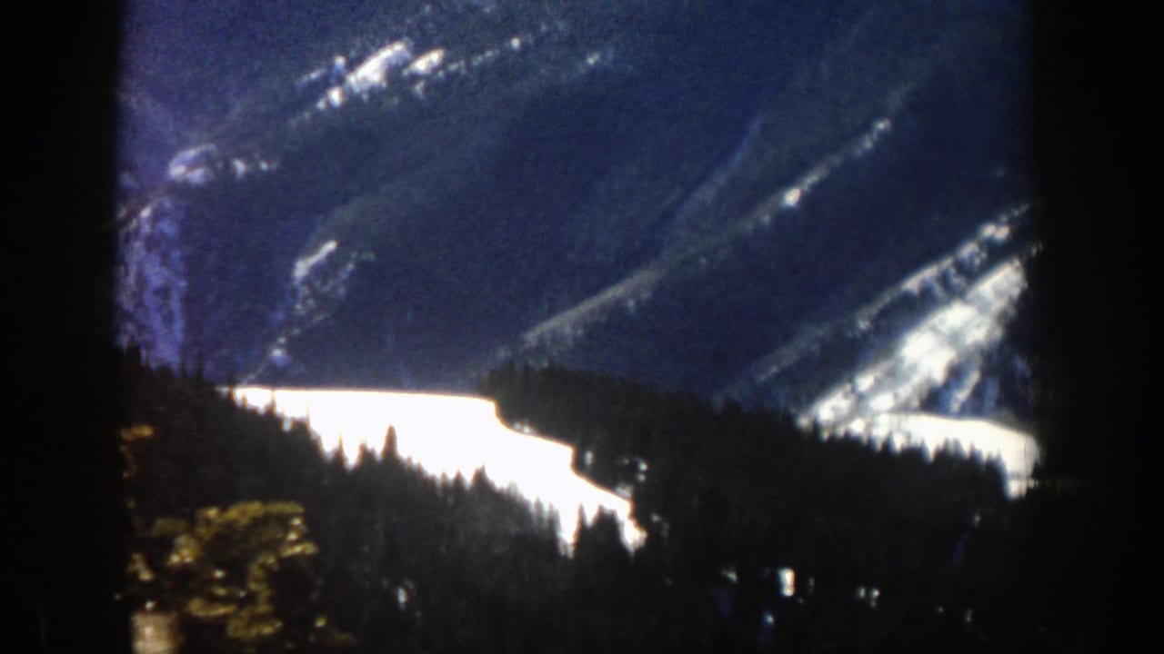 Snowy mountain slope behind a dark evergreen forest, seen through a dark window edge.