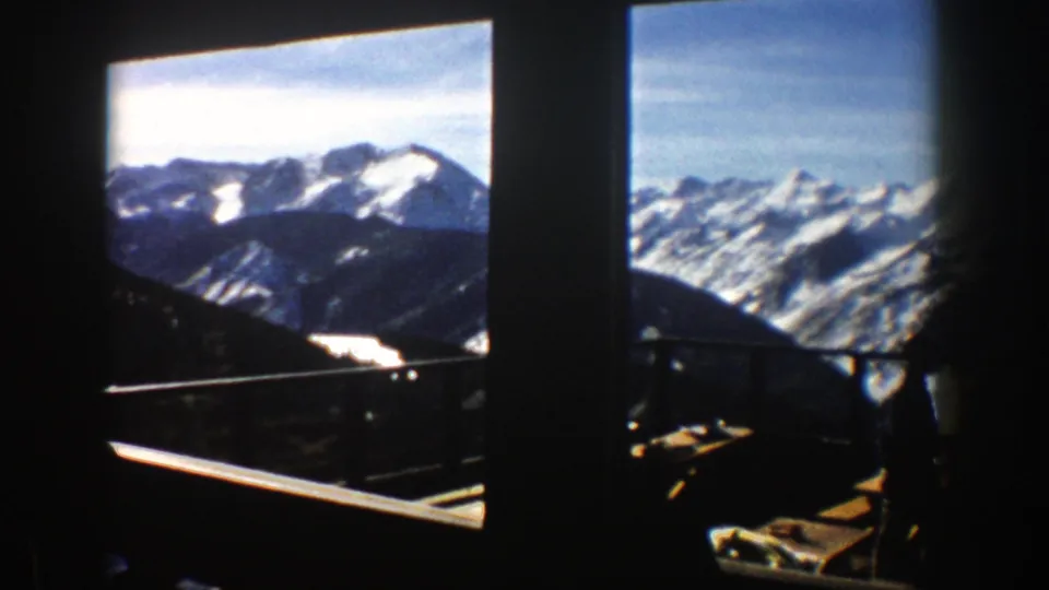 1961 home-movie view from Aspen showing snowy peaks, a balcony rail, and dark interior or window edges.