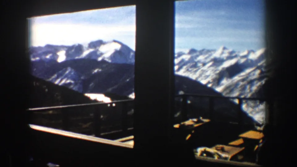 1961 Aspen footage shows a snowy mountain range through broad window openings, with a balcony railing and seating in shadow.