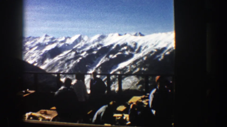 Aspen 1961 home-movie frame with silhouetted men at picnic tables on a balcony overlooking snow-capped mountains.