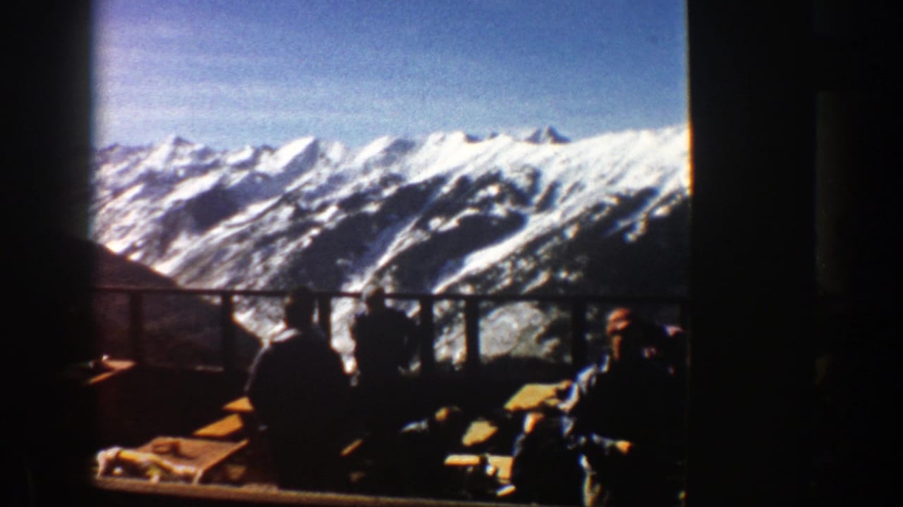 1961 Aspen home-movie frame showing dark silhouettes on a balcony against bright snow-covered mountains and blue sky.