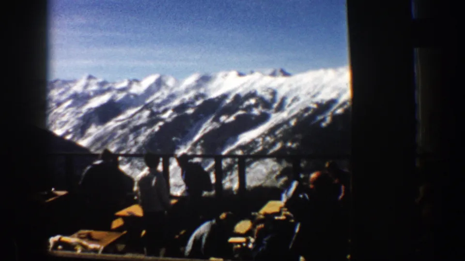 1961 8mm frame of a ski-resort overlook, with silhouetted visitors, balcony furniture, and snow-bright peaks.