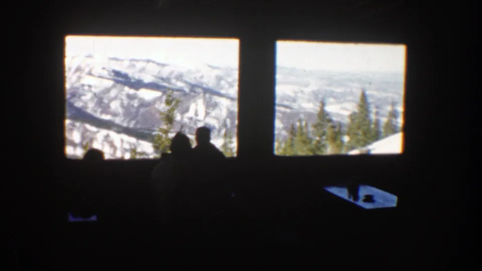 Dark lodge interior with silhouetted figures framed by windows looking out to Aspen snowfields and evergreen trees.