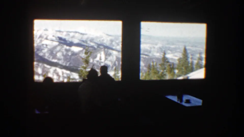 1961 Aspen home-movie frame showing silhouetted people indoors looking through large windows at snow-covered mountains and evergreen trees.