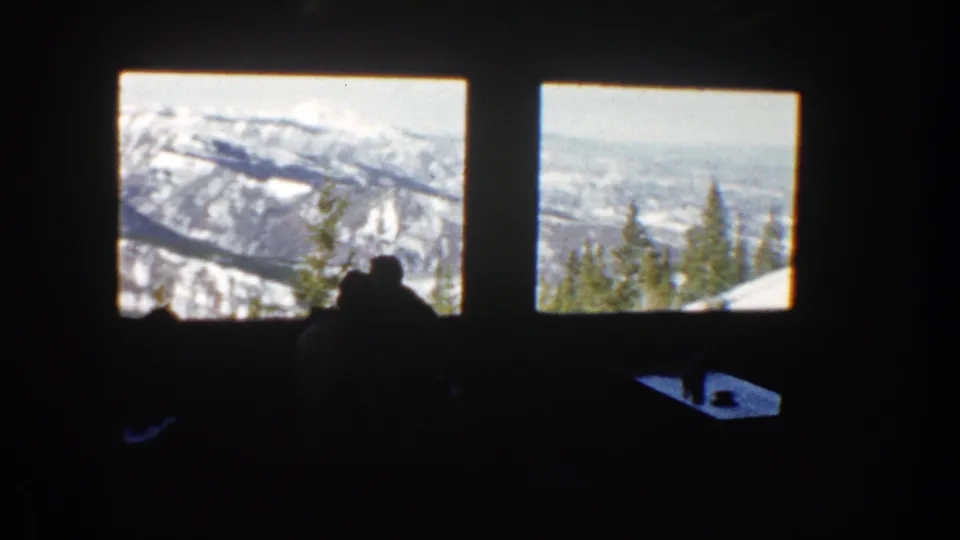 Aspen, Colorado footage frame with people in silhouette inside a lodge-like room, facing two windows and a snowy mountain landscape.