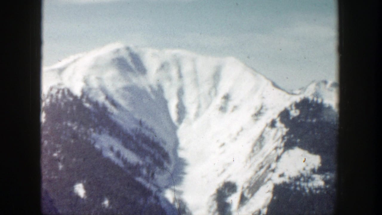 1961 8mm Aspen, Colorado view of a snowy mountain face under blue sky, framed by dark film gate edges.
