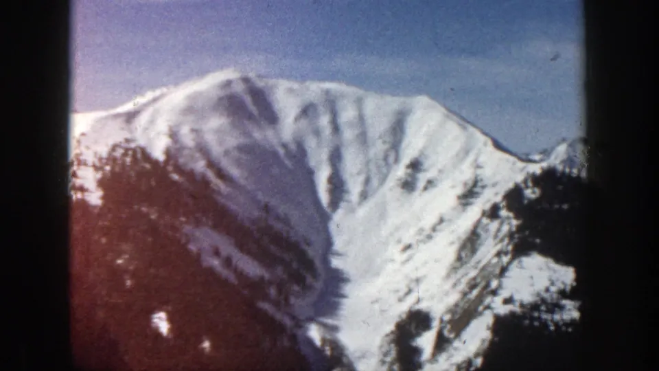 1961 Aspen home-movie frame showing the mountain face more prominently, with snow gullies, trees, and 8mm color variation.