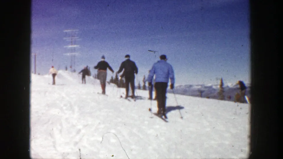 1961 Aspen home-movie view of skiers ascending a snowy slope, including a figure in a blue jacket and distant mountain ridges.