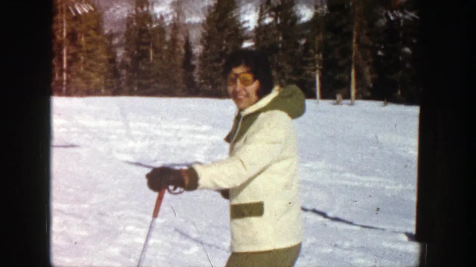 1961 Aspen home-movie frame showing a skier in a cream jacket and goggles holding poles against snowy ground and trees.