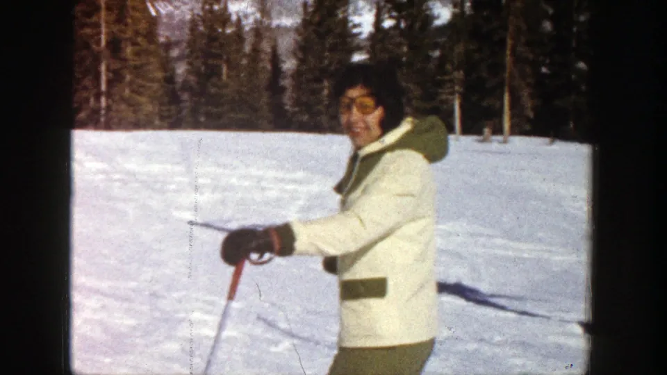 1961 Aspen frame with a smiling skier in winter gear holding ski poles, with snow and evergreen trees behind.