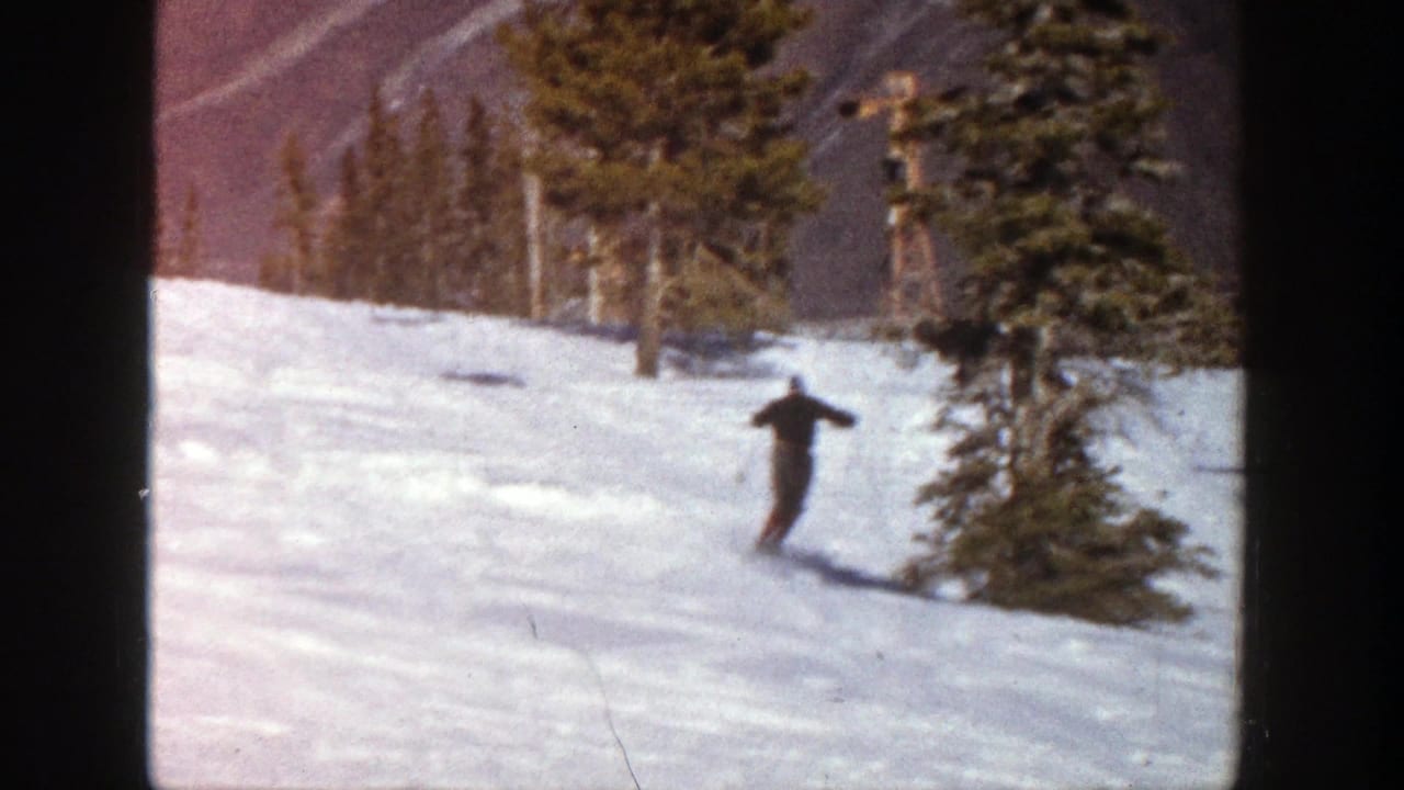 In 1961 Aspen, Colorado footage, an adult skier in dark clothing moves downhill across snow with pines and mountains behind.