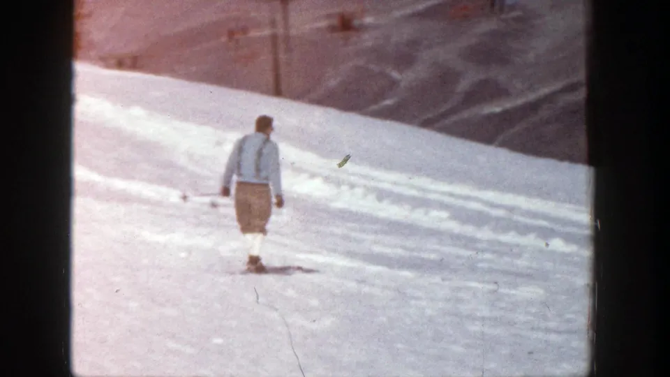 An adult skier in a light blue shirt and suspenders skis away on the snowy Aspen slope, holding poles in the 1961 home movie frame.