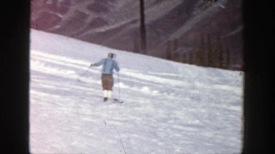 The light-clothed adult skier continues down the Aspen snow slope, with distant trees, mountain terrain, and handheld 8mm framing visible.