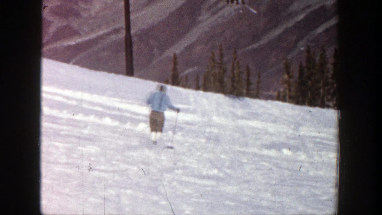 Skier in a light jacket moving across a snowy slope with trees and mountains behind.