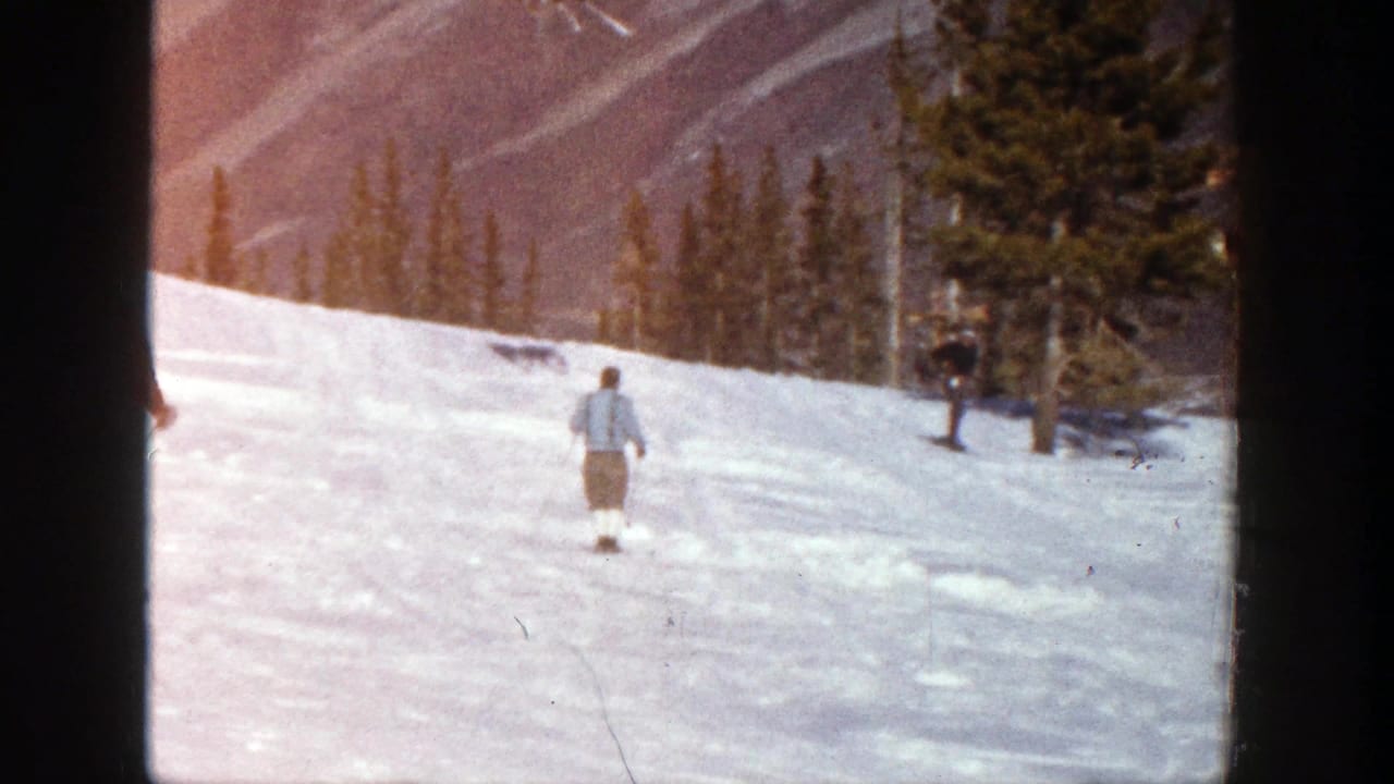 Distant skier in light clothing on a snowy slope near pine trees and mountain ridges.