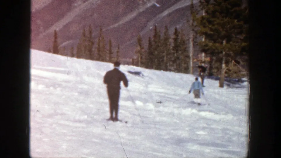 1961 Aspen home-movie frame showing two skiers on the slope, with one nearer the camera and another moving toward the trees.