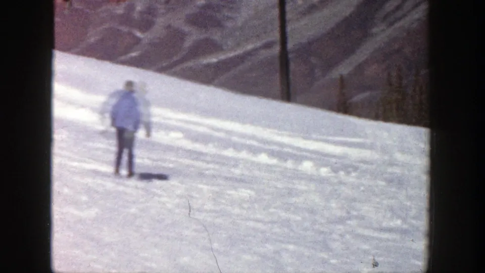1961 Aspen frame of a skier viewed from behind on a sunlit snowy slope, with pine trees and mountain ridges beyond.
